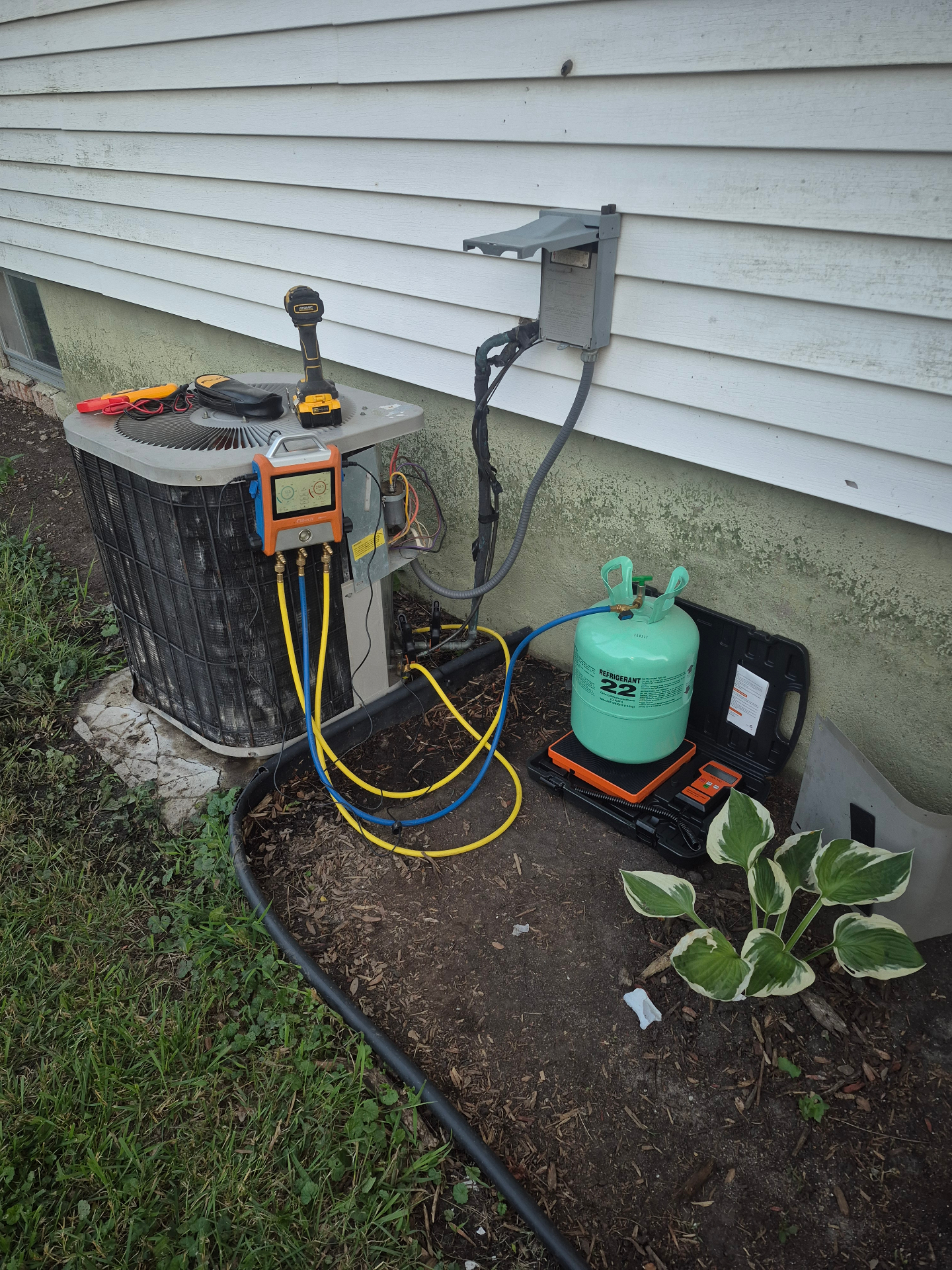 Outdoor AC condenser hooked up to digital manifold gauges and R-22 refrigerant tank on a scale during a recharge