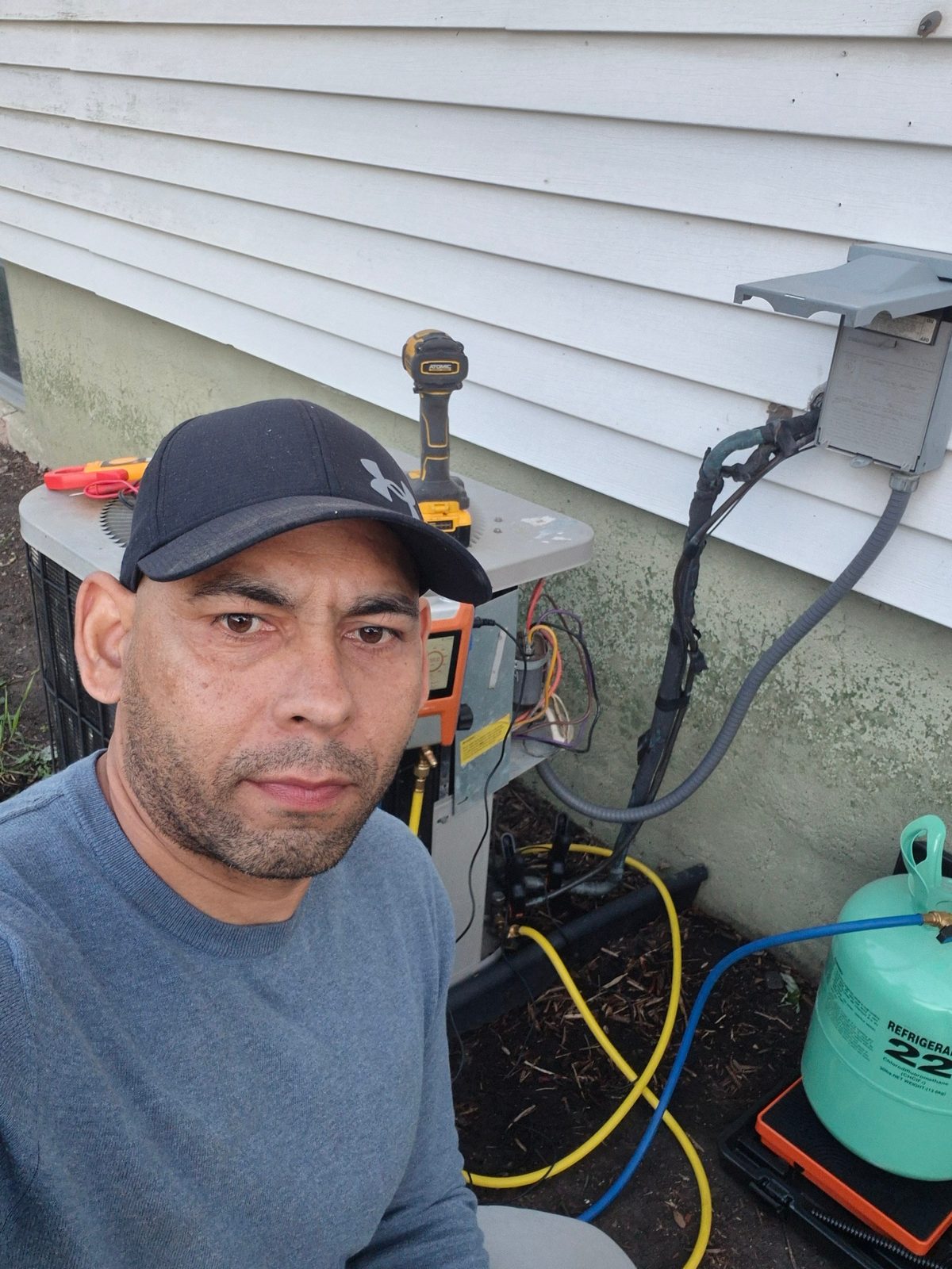 Gerardo HVAC technician on site next to an outdoor AC unit with manifold gauges, drill and refrigerant tank during a service call